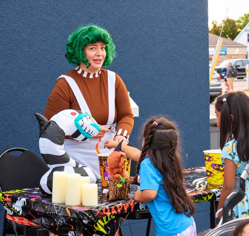 A green-wigged costumed host chats with a child beside an activity table.