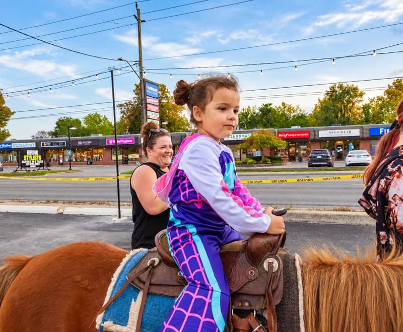 A child in a colourful costume rides a small pony beside an adult handler.