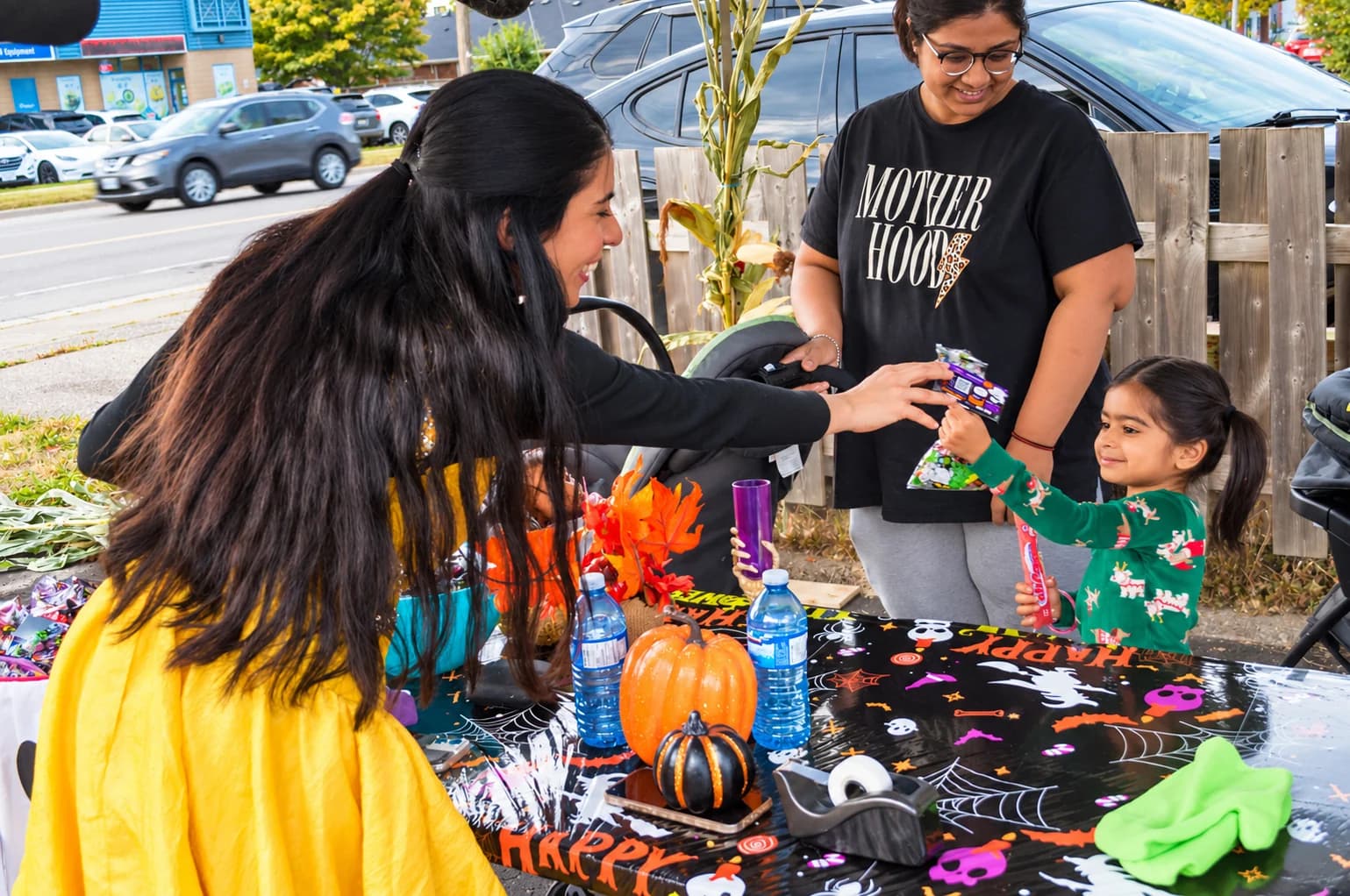 A host in black and yellow hands a gift to a child in green at a Halloween activity table.