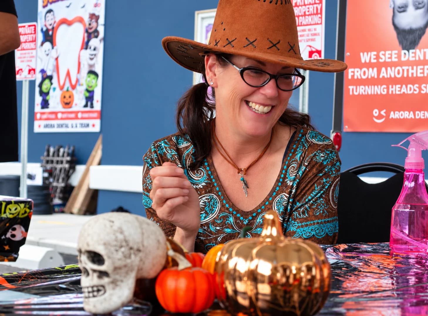 A smiling woman in a brown cowboy hat sits behind a table of Halloween decorations.